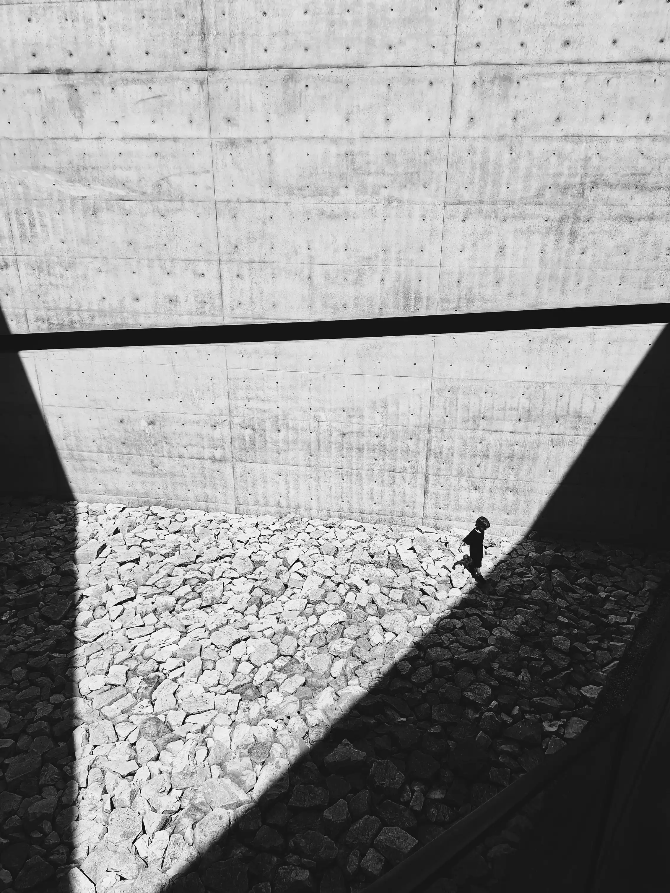 Parede de betão à vista com sombra diagonal sobre brita branca e uma figura solitária