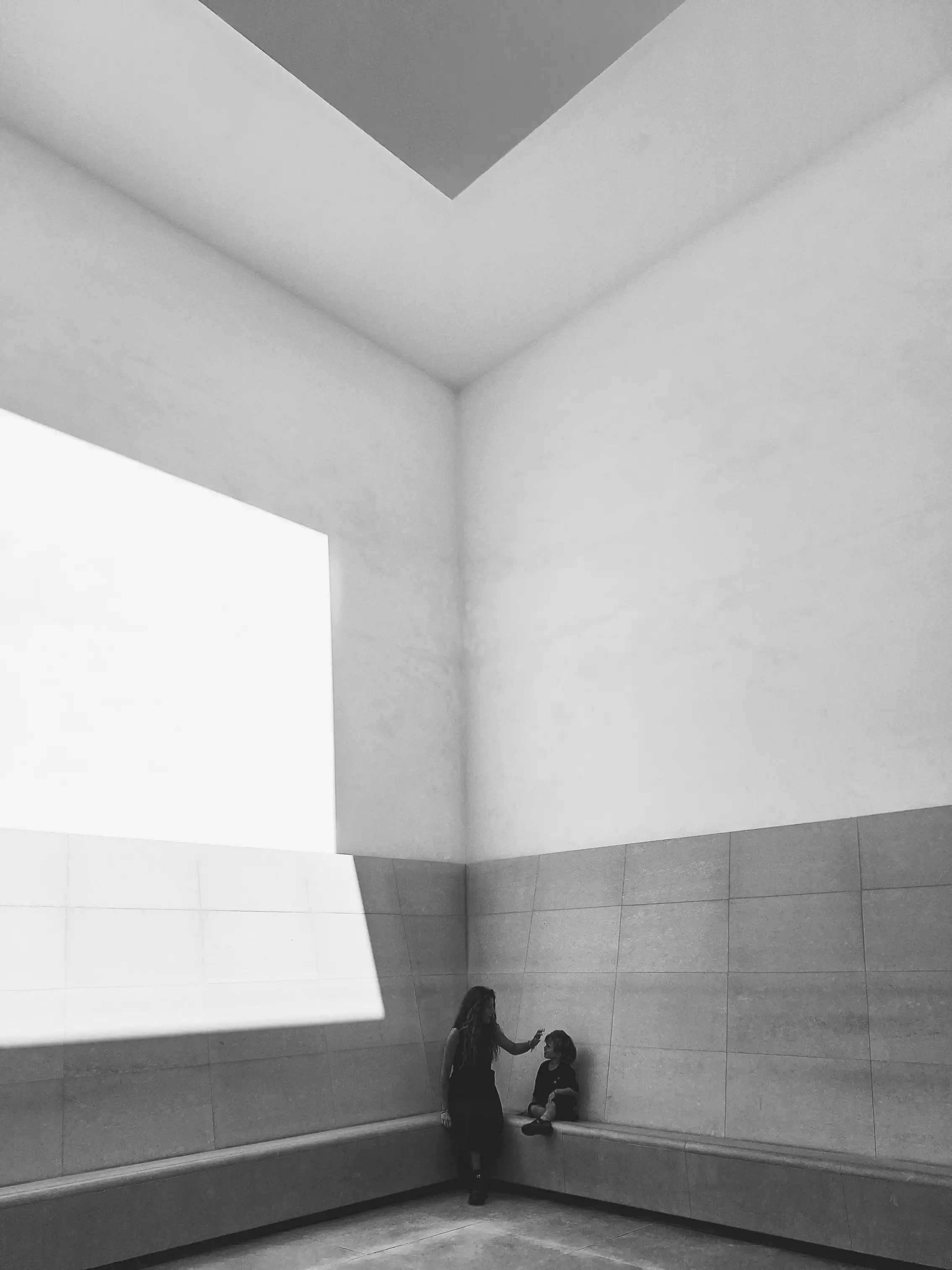 Interior amplo com clarabóia angular; uma mulher junto a um banco de pedra estende a mão a uma criança